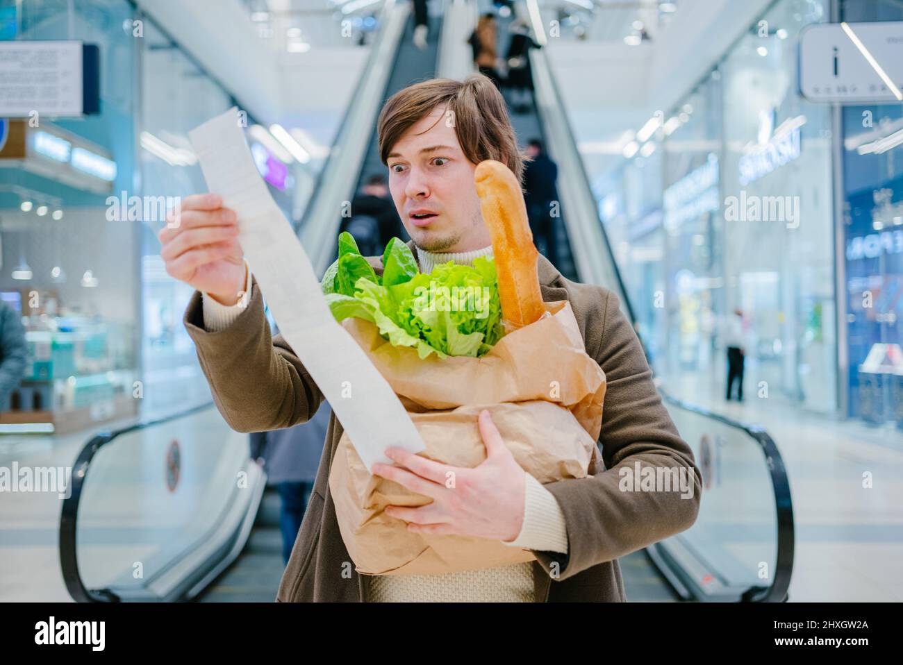 L'homme en manteau tient un chèque en papier et un sac avec une baguette et de la laitue dans un centre commercial et est choqué par les prix élevés des produits d'épicerie. Banque D'Images