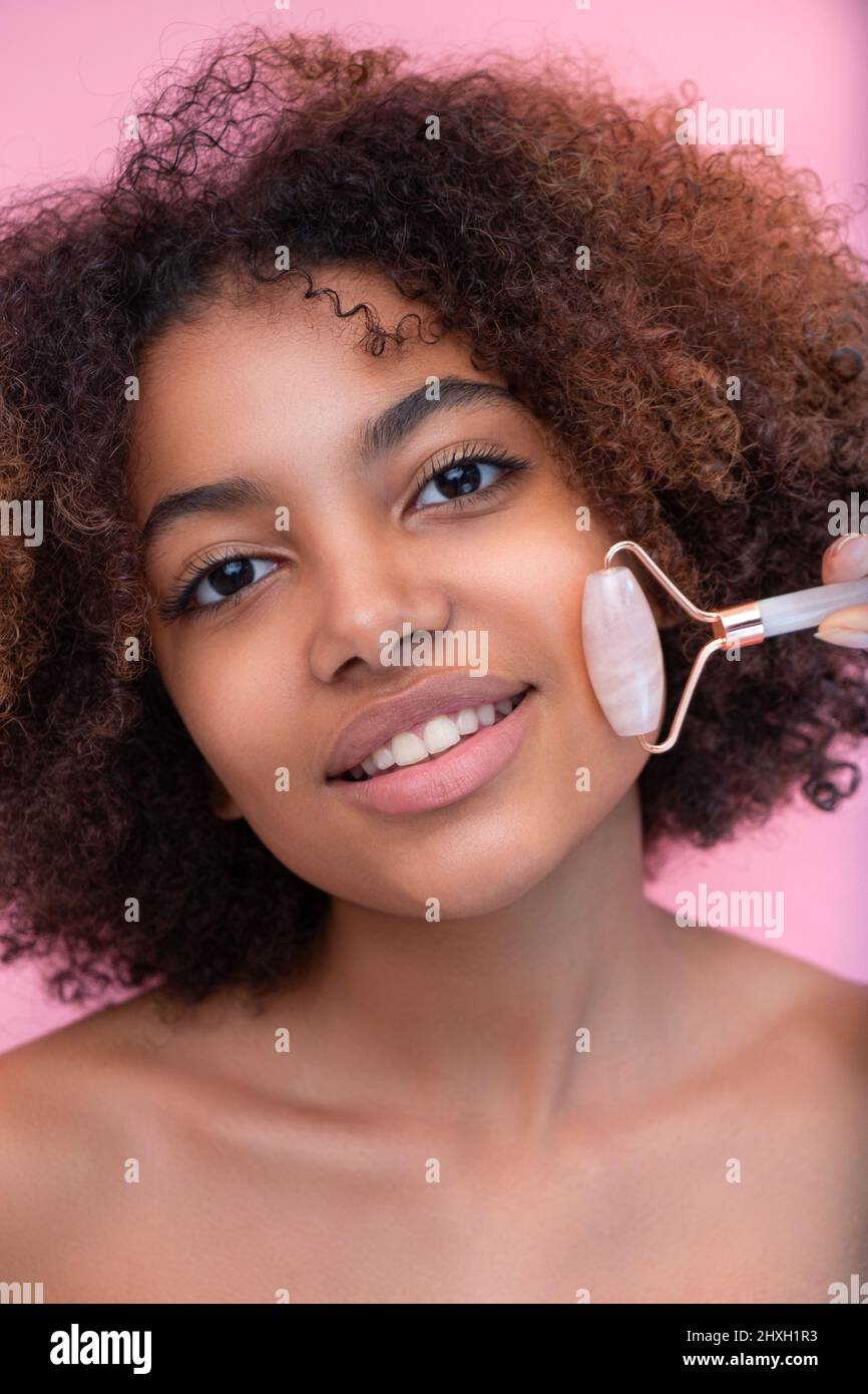 Portrait d'une jeune femme à la peau sombre et bien entretenue avec des cheveux luxuriants, faisant un massage du visage avec l'aide d'un rouleau avec les yeux fermés sur un fond rose gros plan, les soins et les soins de la peau Banque D'Images