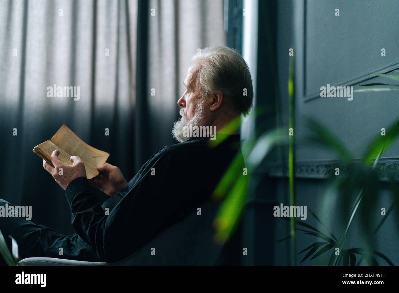Vue latérale d'un homme âgé aux cheveux gris concentré lisant un livre à la maison assis à table, détendez-vous et reposez-vous avec de la littérature. Banque D'Images