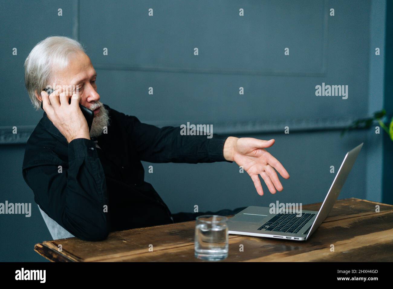 Vue de face d'un homme âgé aux cheveux gris en colère qui parle à l'assistance technique et demande de l'aide pour réparer un ordinateur portable cassé assis à une table en bois. Banque D'Images