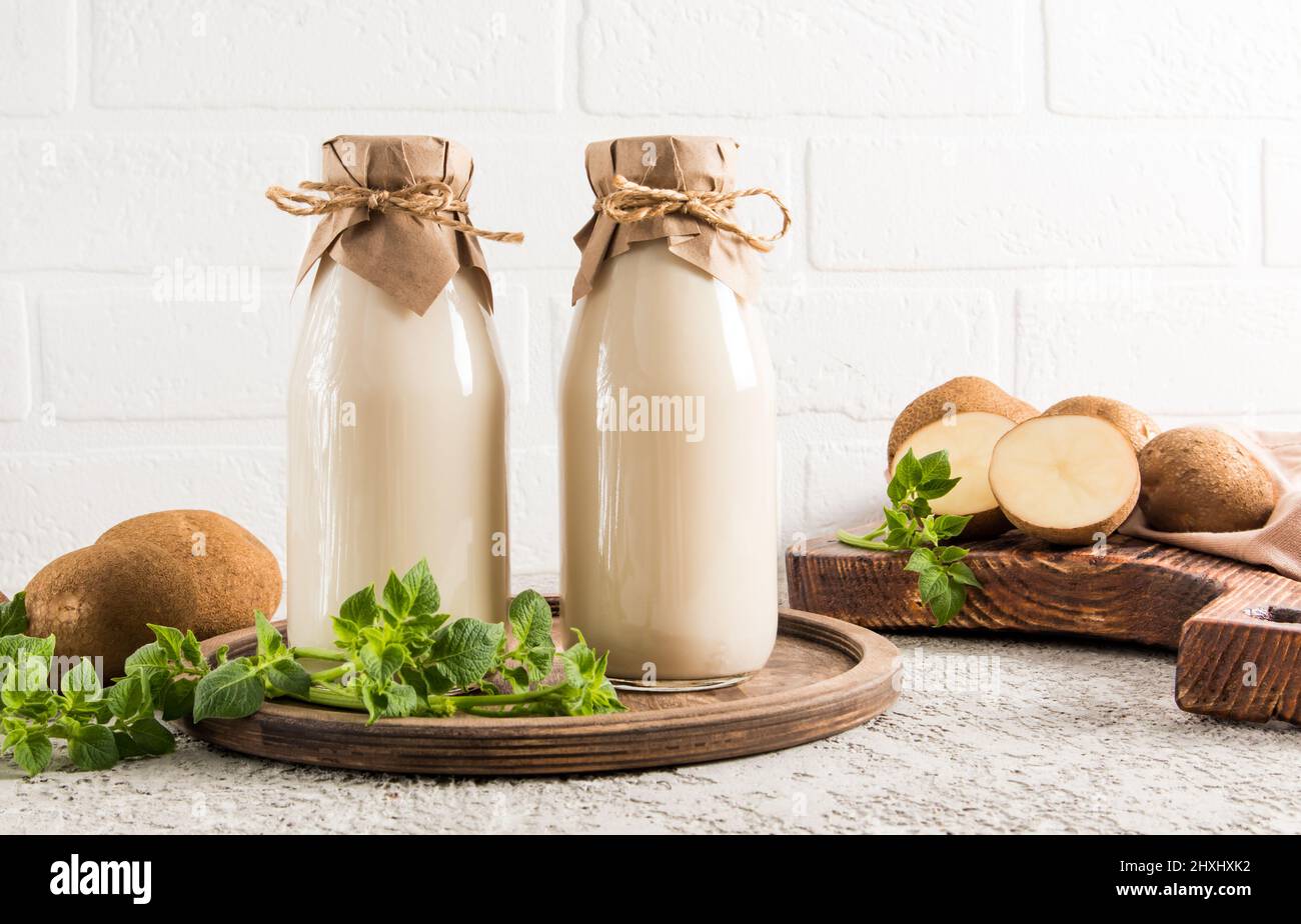 deux bouteilles de lait de pomme de terre à base de plantes sur un plateau rond en bois face d'un mur de briques blanches. tubercules de pomme de terre hachés sur une planche. lait de remplacement Banque D'Images