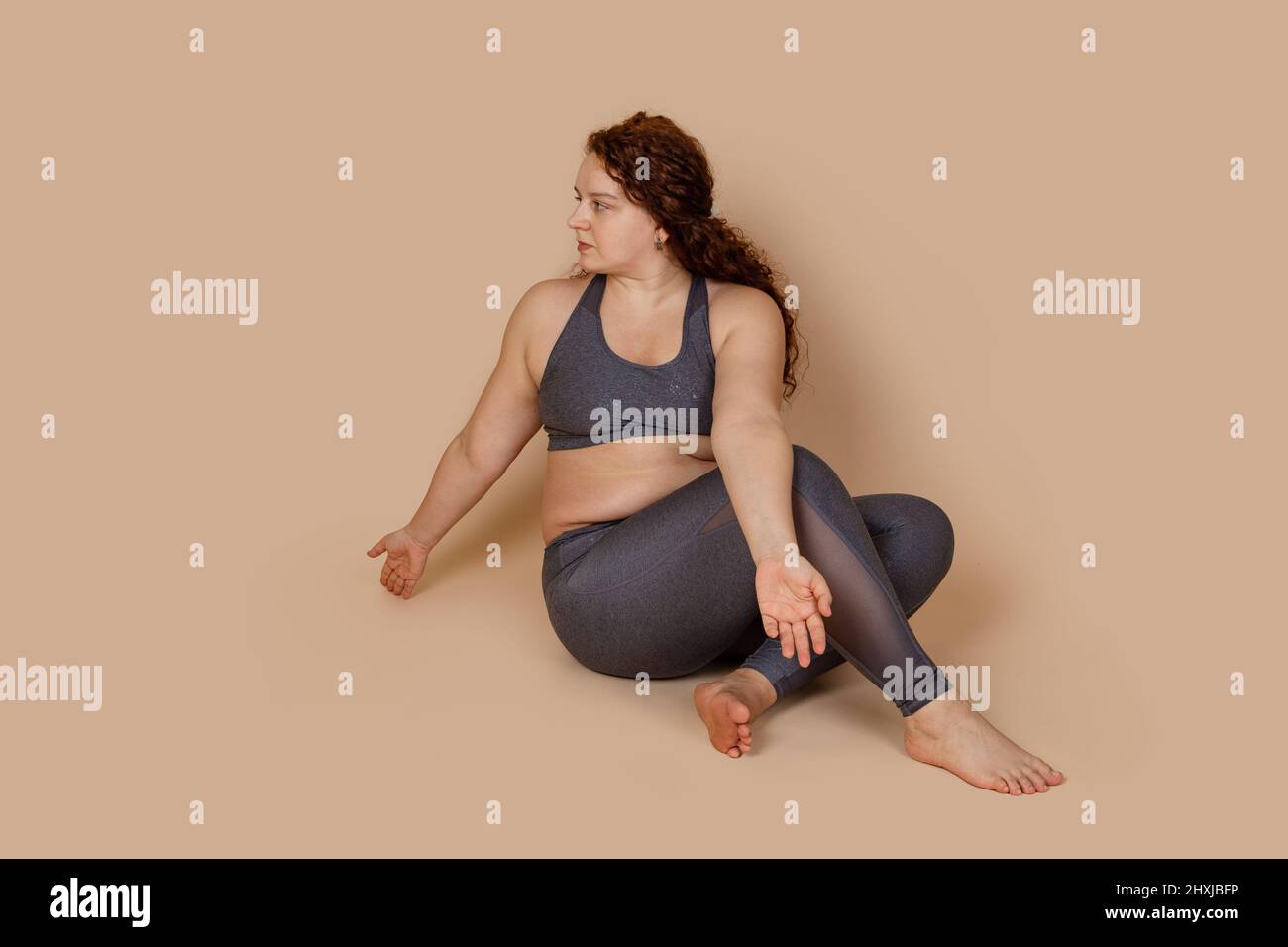 Femme au gingembre aux cheveux bouclés assise sur le sol, en regardant de côté avec des positions de bras ouvertes, à mi-tour. La forme physique gagne du poids Banque D'Images