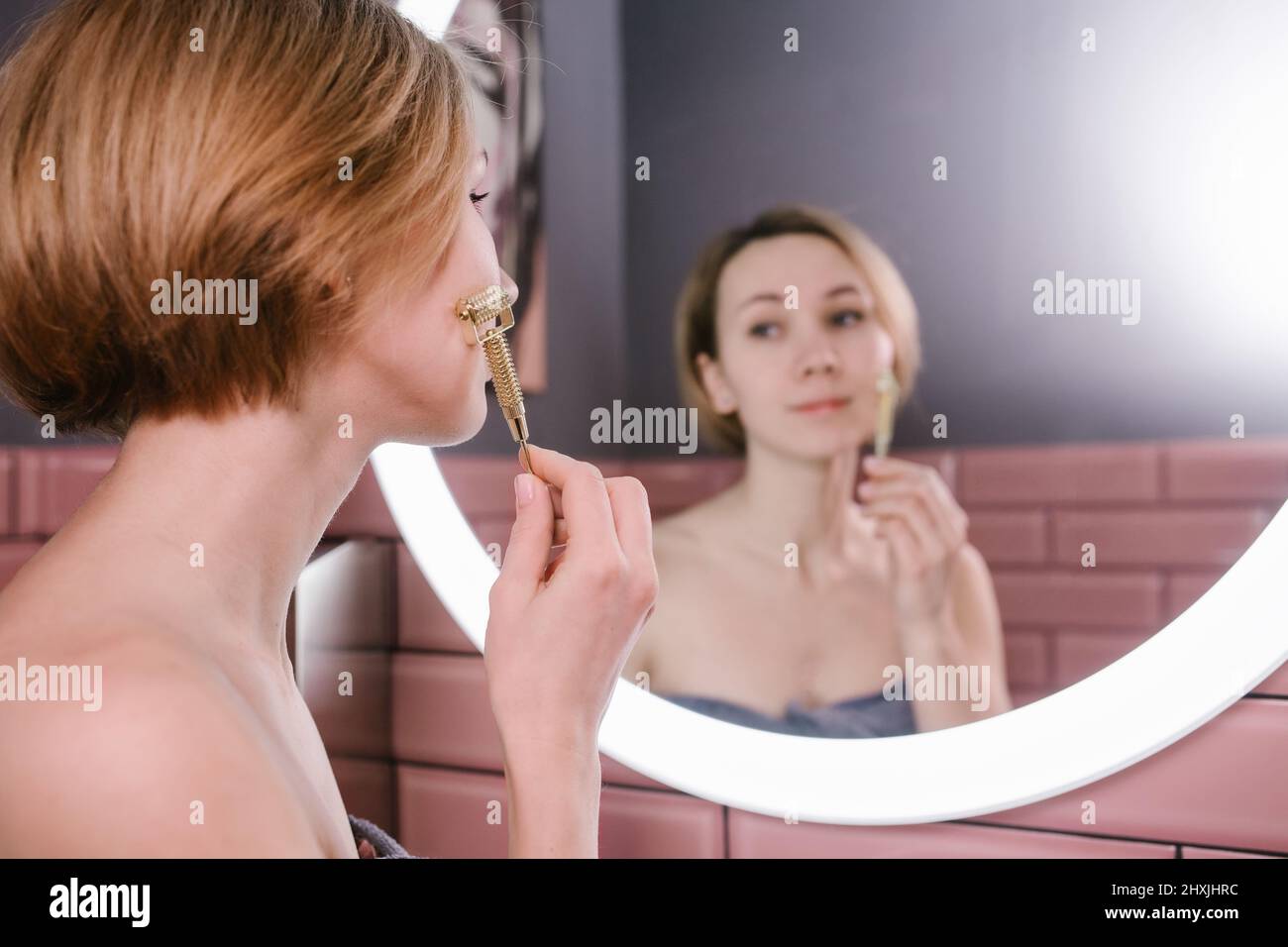 Femme à l'élévation massante visage avec rouleau de gua sha devant un miroir de salle de bains Banque D'Images