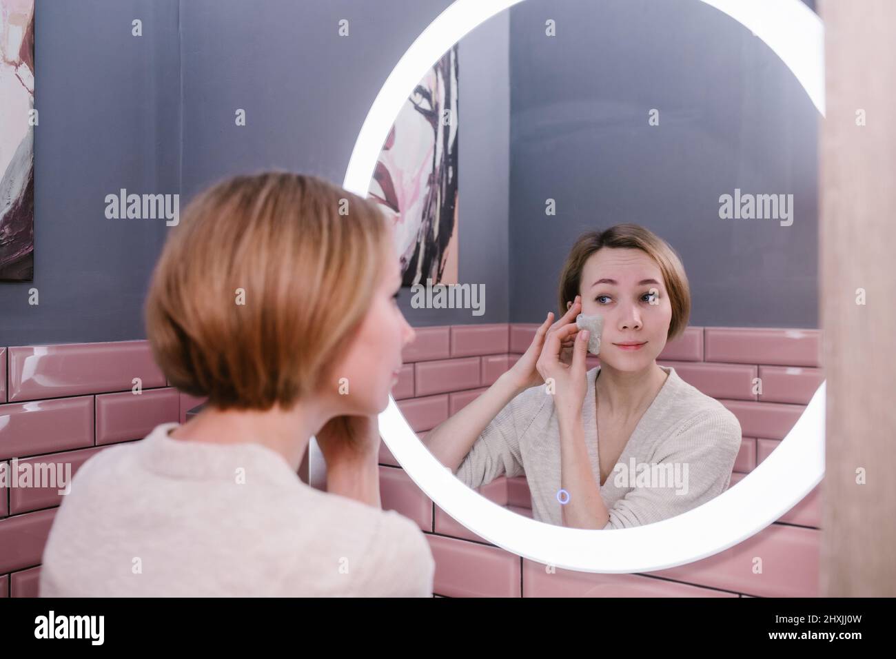 Bonne femme massant son visage avec de la pierre de gua sha devant un miroir de salle de bains Banque D'Images