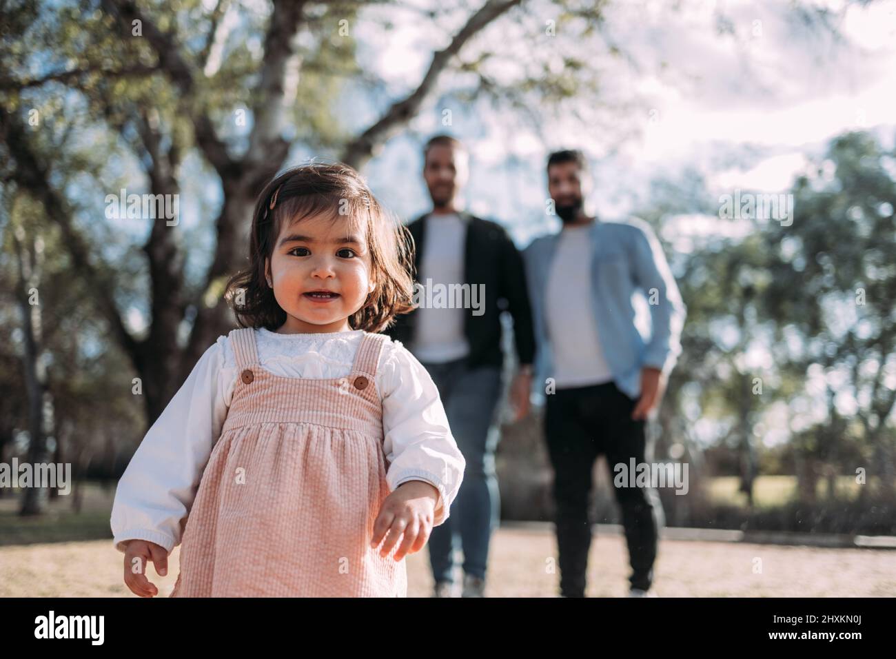 Une petite fille s'approche curieusement de la caméra avec ses parents la regardant en arrière-plan. Famille moderne. Banque D'Images