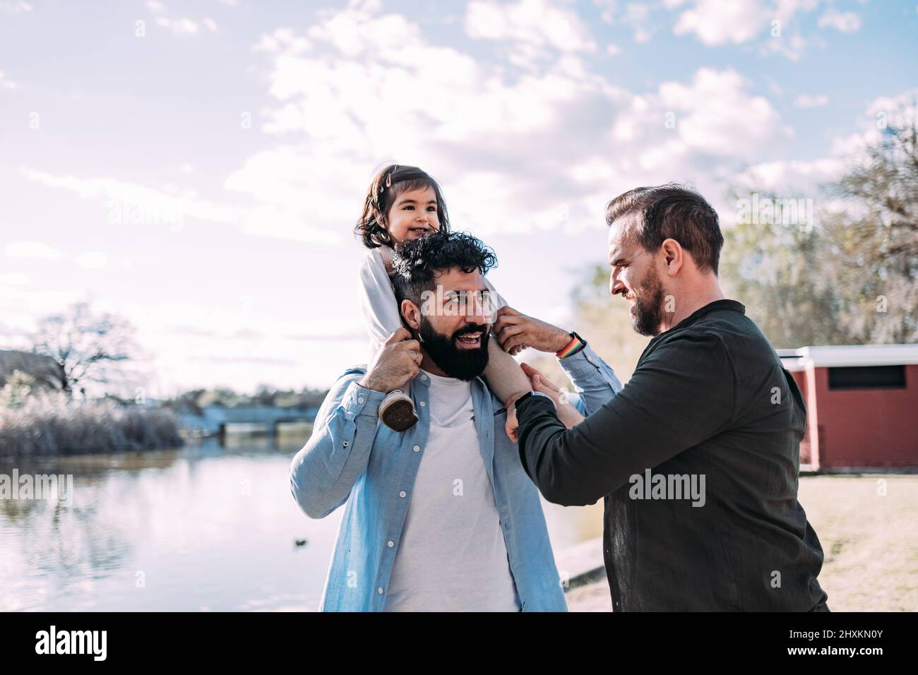 Couple gay avec leur petite fille sur la rive d'un lac dans le parc. Le père tient la fille sur ses épaules piggyback. Banque D'Images