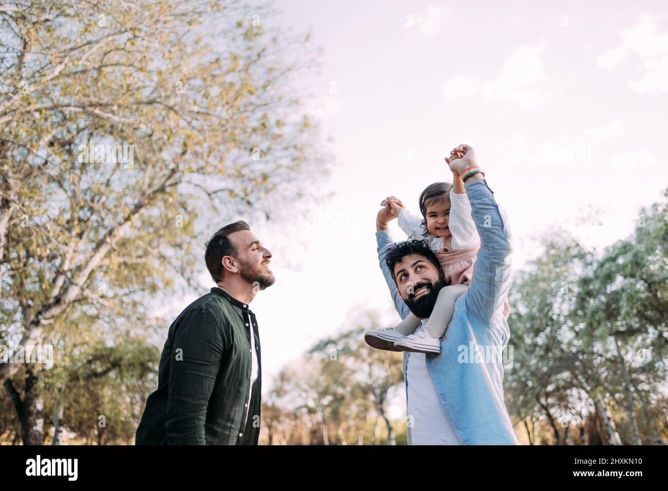 Couple gay avec leur petite fille dans le parc. Le père tient la fille sur ses épaules dans une promenade de pigeyback et ils élèvent leurs bras heureusement. Banque D'Images