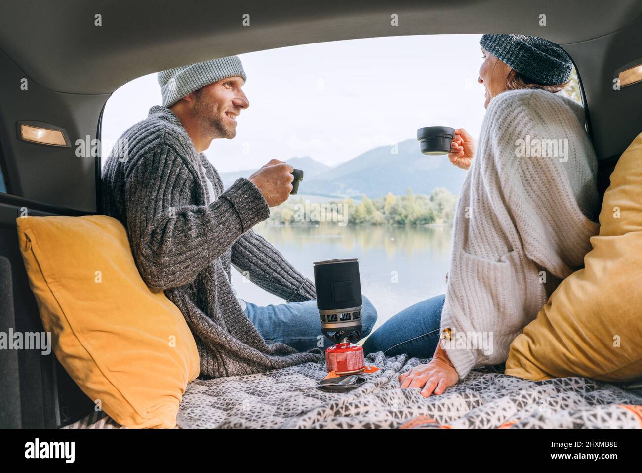 Coffre de voiture vue de chat couple habillé chaud vêtements tricotés appréciant cuisinière à gaz café préparé et vue sur le lac de montagne. Confortable début automne couple aut Banque D'Images