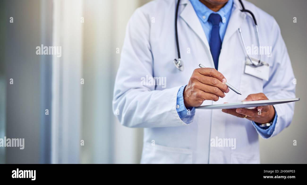 Ses horaires sont toujours occupés. Prise de vue d'un médecin masculin méconnu qui écrit sur une tablette numérique tout en se tenant à l'intérieur d'un hôpital pendant la journée. Banque D'Images