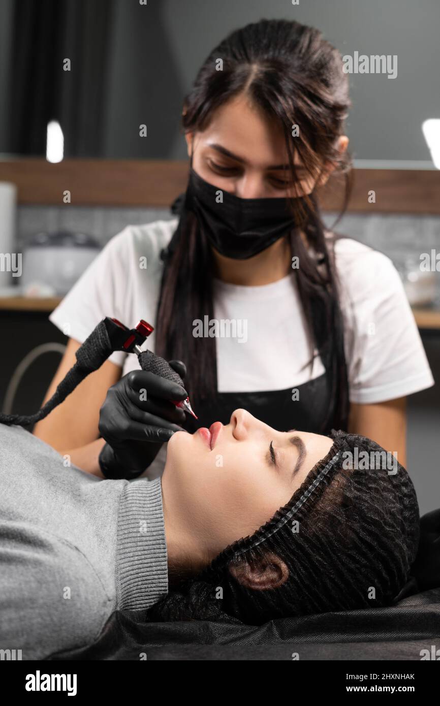 Femme tatouant permanent maquillage de lèvre dans le salon de beauté, augmentation de lèvre par application de pigment, cosmétologue faisant la procédure de maquillage en utilisant la machine de tatouage Banque D'Images