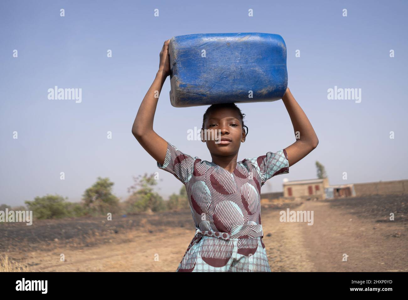 Fière jeune fille africaine qui a l'air de marcher depuis la maison isolée de ses parents jusqu'au village lointain, portant une bouteille d'eau sur sa tête ; le droit de l'homme à Banque D'Images