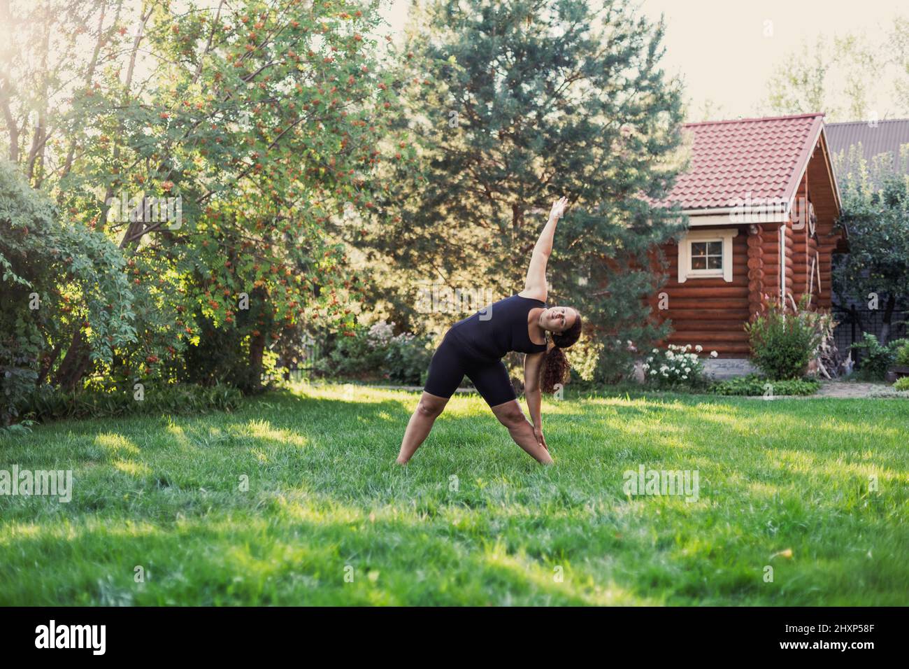 Heureuse femme sur-pondérée faisant des exercices de yoga debout sur le sol étirant le corps sur l'arrière-cour de la maison de bois et les arbres en arrière-plan Banque D'Images