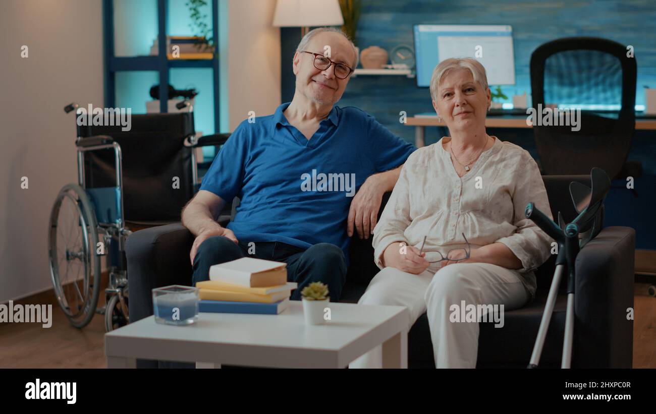 Portrait d'un couple à la retraite avec un handicap assis sur un canapé, regardant la caméra. Homme et femme plus âgés à côté des béquilles et fauteuil roulant pour l'accessibilité et la mobilité, profitant de temps libre. Banque D'Images