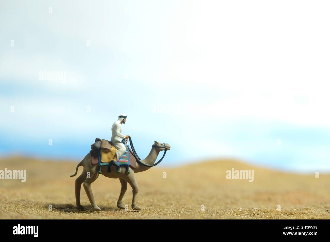 Photographie miniature de personnage de jouet de personnes. Un homme portant des vêtements kandura blancs à cheval à dos de chameau dans un désert dans un ciel lumineux. Photo d'image Banque D'Images