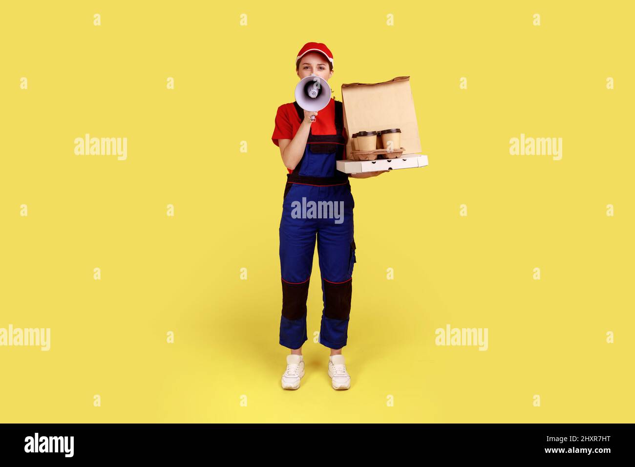 Portrait de la femme de livraison tenant le café avec la pizza, commander la restauration rapide, annonçant la publicité, portant une combinaison et une casquette rouge. Studio d'intérieur isolé sur fond jaune. Banque D'Images