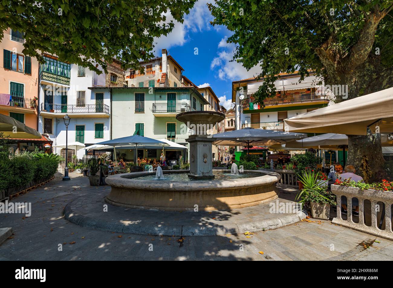 Fontaine et restaurants en plein air sous les arbres sur la petite place de Dolceacqua, Italie. Banque D'Images