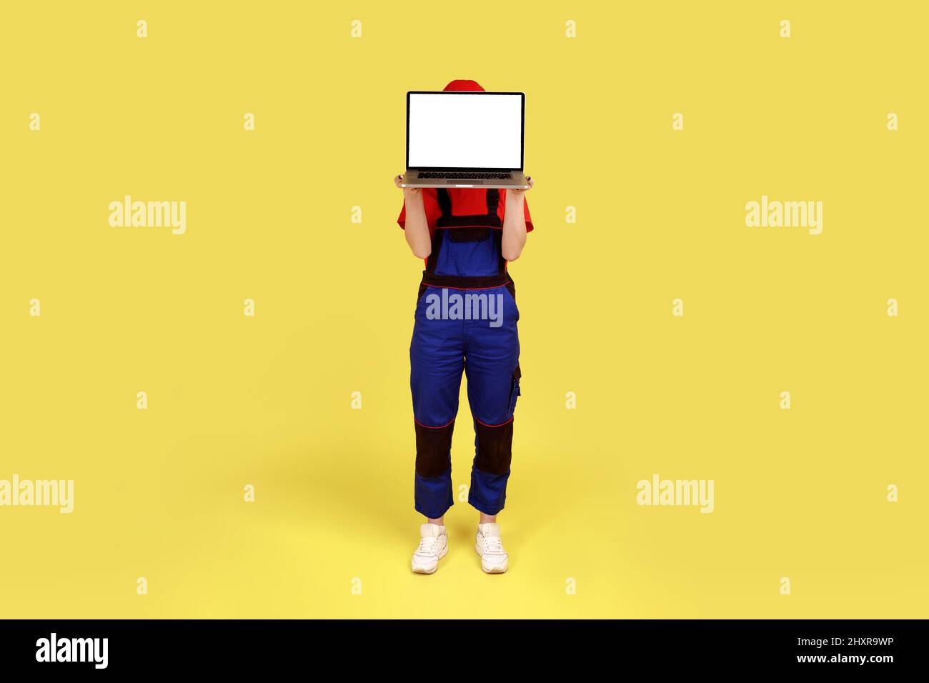Portrait en longueur de la femme employée anonyme debout se cachant derrière un ordinateur portable avec écran vide pour le texte promotionnel, portant un uniforme. Studio d'intérieur isolé sur fond jaune. Banque D'Images