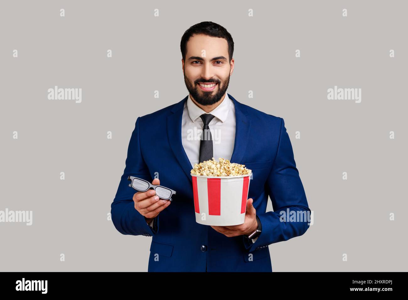 Homme d'affaires à barbe souriant tenant 3D verres et pop-corn, prêt à regarder un film intéressant, portant un costume de style officiel. Prise de vue en studio isolée sur fond gris. Banque D'Images