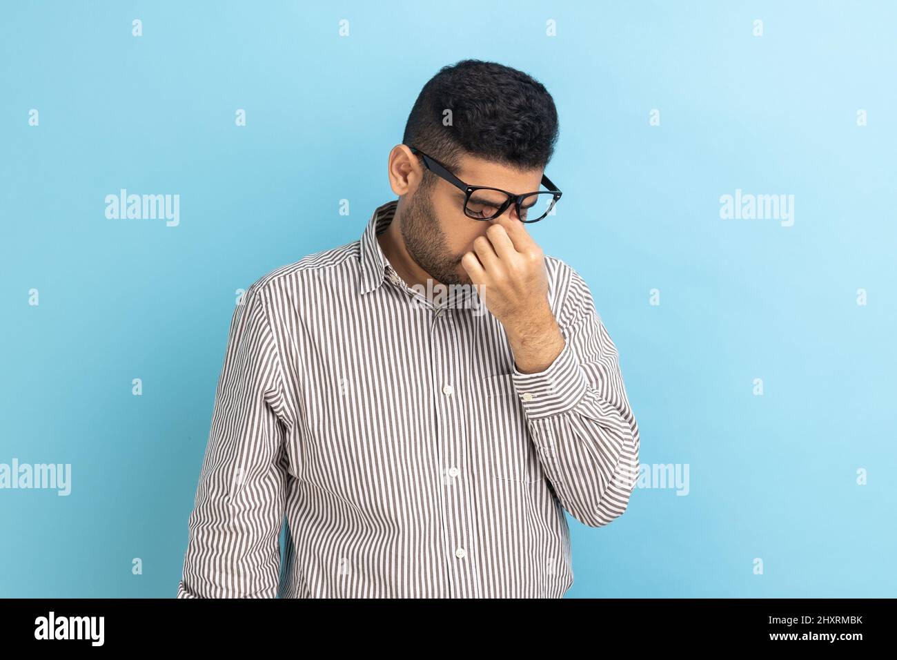 Portrait d'un homme d'affaires frustré dans des lunettes se frottant les yeux, étant fatigué de longues heures de travail sur ordinateur portable, douleur ou douleur, portant une chemise rayée. Studio d'intérieur isolé sur fond bleu. Banque D'Images