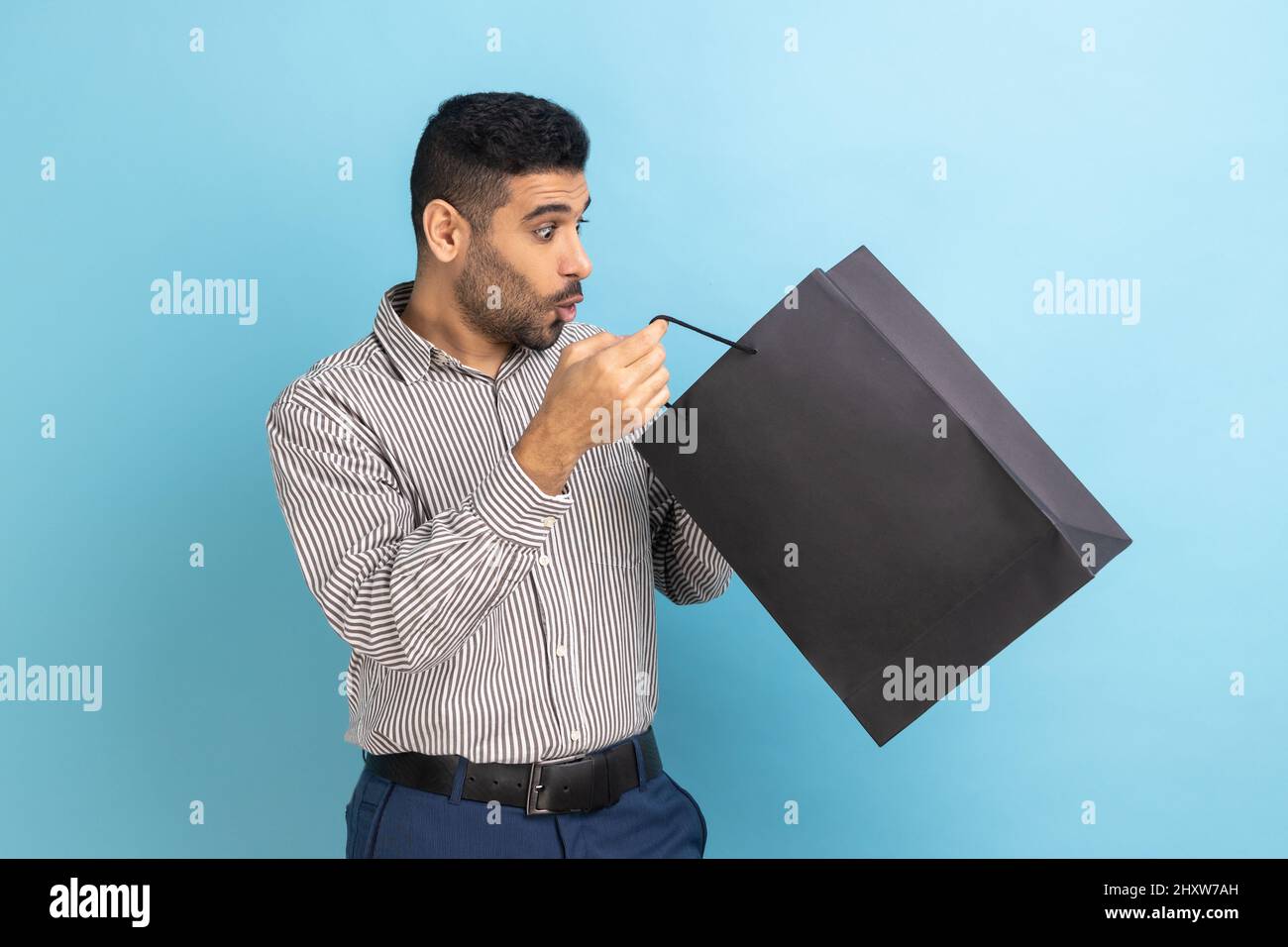 Surprise homme d'affaires barbu regardant à l'intérieur de sac de shopping avec les yeux grands ouverts la bouche dans étonnement, choqué par l'achat, portant une chemise rayée. Studio d'intérieur isolé sur fond bleu. Banque D'Images