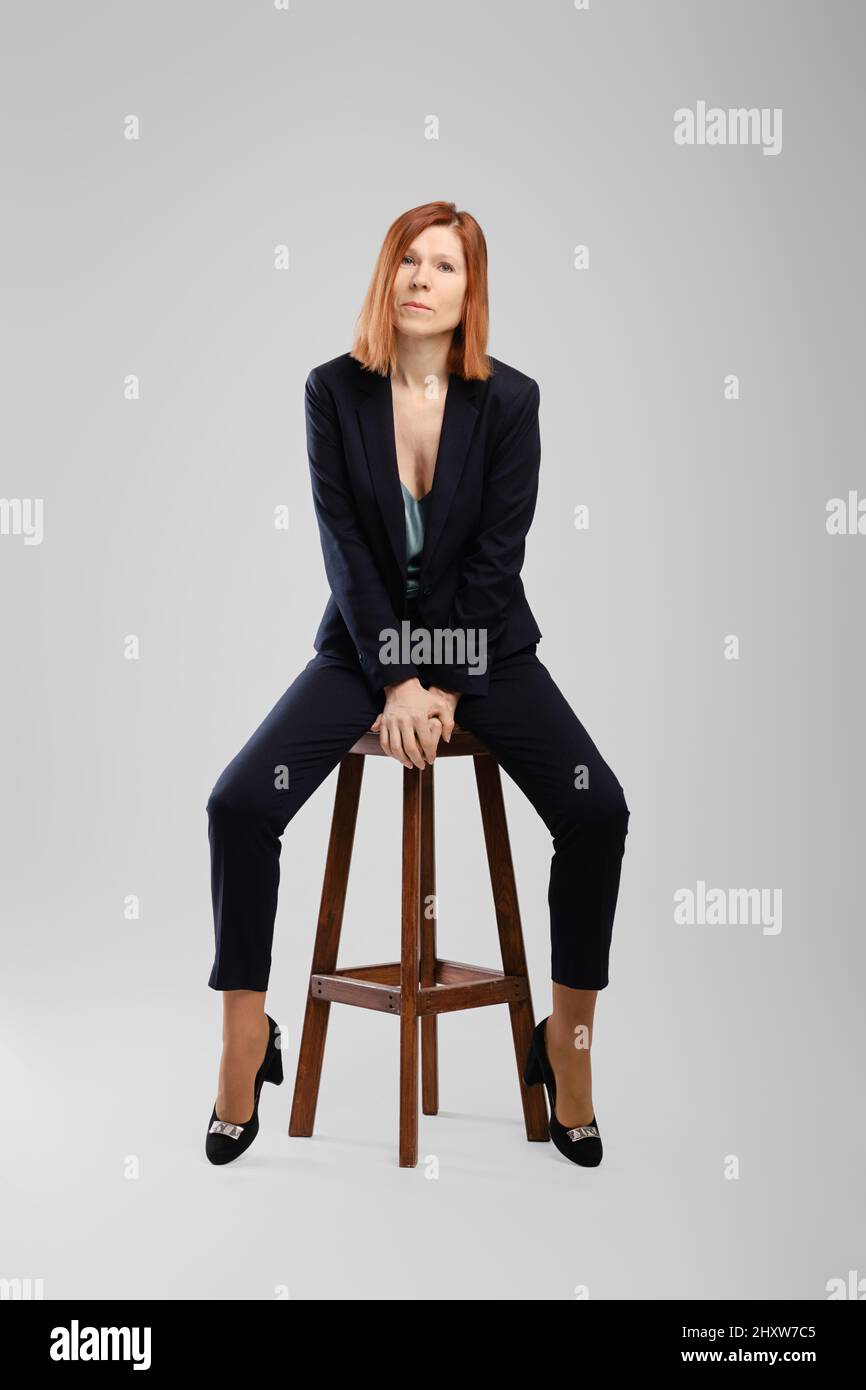 Élégante femme d'âge moyen en costume-culotte posé en studio assis sur une chaise avec des jambes étalées sur fond gris Banque D'Images