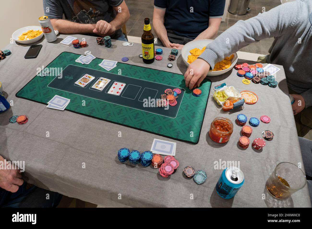 Des amis masculins jouant au Texas Hol 'em Poker sur une table à la maison avec des jetons de jeu, un tapis de poker, des jetons et de la sauce salsa et des bières. Angleterre Banque D'Images