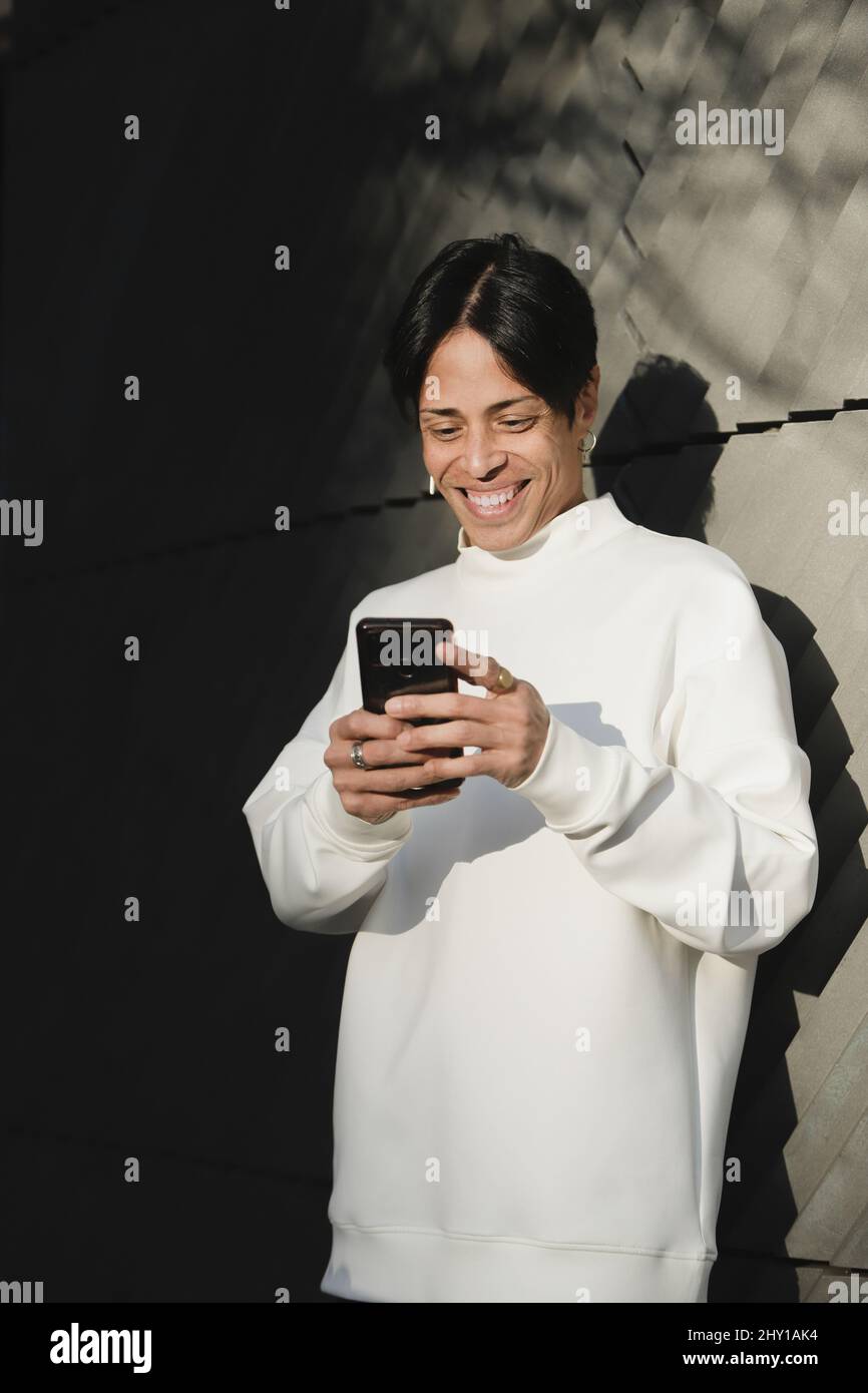 Homme asiatique souriant en pull blanc debout sur un mur en béton de rue et lisant des messages sur un smartphone Banque D'Images