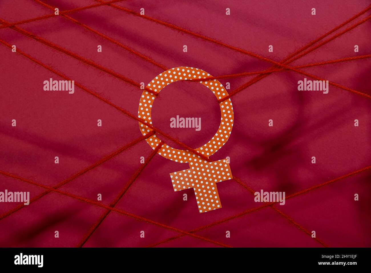 Le symbole de Vénus à point de polka orange est attaché avec des cordes représentant l'inégalité des sexes dans la pièce avec de l'ombre et un éclairage tamisé sur fond rouge Banque D'Images