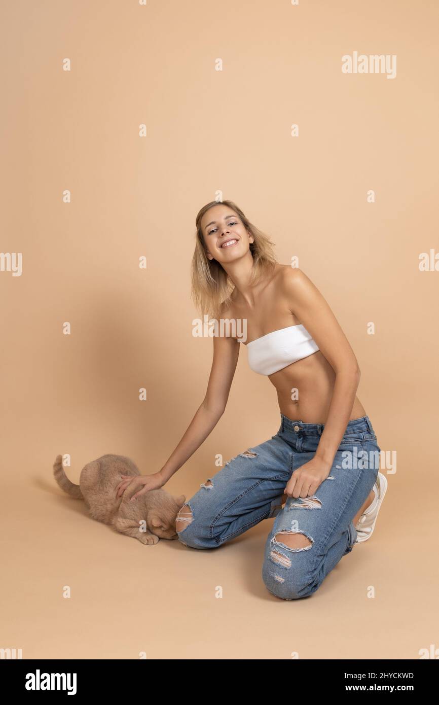 Portrait d'une femme portant un Jean déchiré souriant petting pêche chat de couleur avec la main regardant l'appareil photo sur fond de couleur de peau. Personne jouant avec Banque D'Images