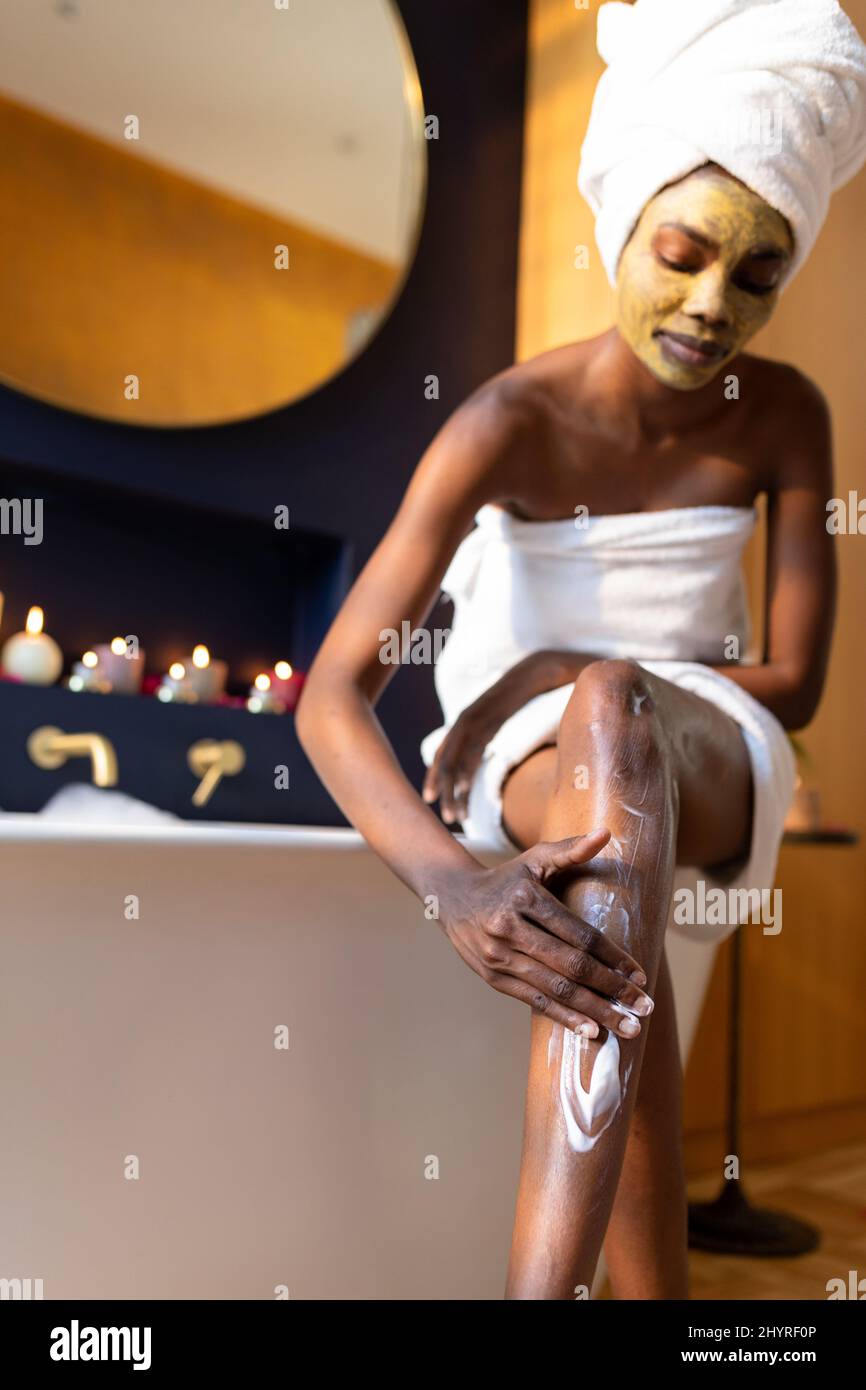Femme afro-américaine avec masque de visage appliquant de la crème sur la jambe dans la salle de bains à la maison Banque D'Images