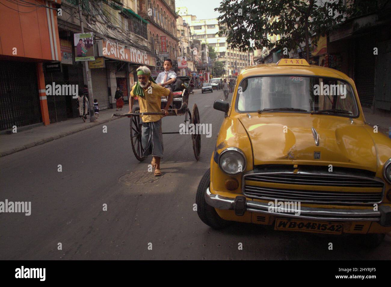 Un arrache-pousse transportant un enfant pour l'école, se déplaçant près d'un taxi jaune qui est garée dans la rue de Kolkata, Bengale-Occidental, Inde. Au fil des ans, les groupes de défense des droits de l'homme et les autorités gouvernementales ont essayé de limiter l'utilisation de pousse-pousse tirés à la main, selon Matters India dans une publication de juillet 2021. Le pousse-pousse tiré à la main aurait été considéré comme un « anachronisme colonial dégradant », a déclaré le rapport, ajoutant que « les autorités locales ont officiellement interdit les véhicules en 2006 et ont cessé de délivrer ou de renouveler les licences ». Banque D'Images