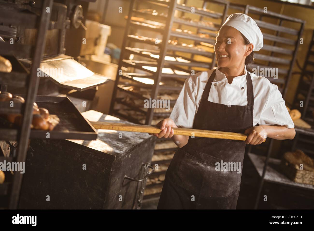 Galette asiatique mi-longue pour adulte souriante gardant un plateau à pain tressé avec une longue spatule dans le rack Banque D'Images