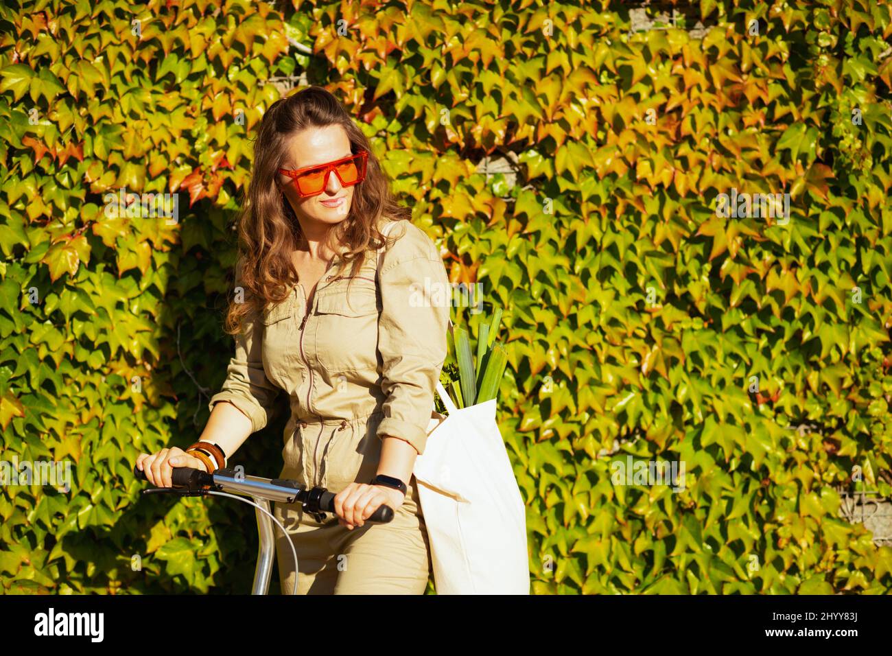 élégante femme de 40 ans en lunettes de soleil et combinaison avec sac fourre-tout et scooter contre le mur vert en plein air dans la ville. Banque D'Images