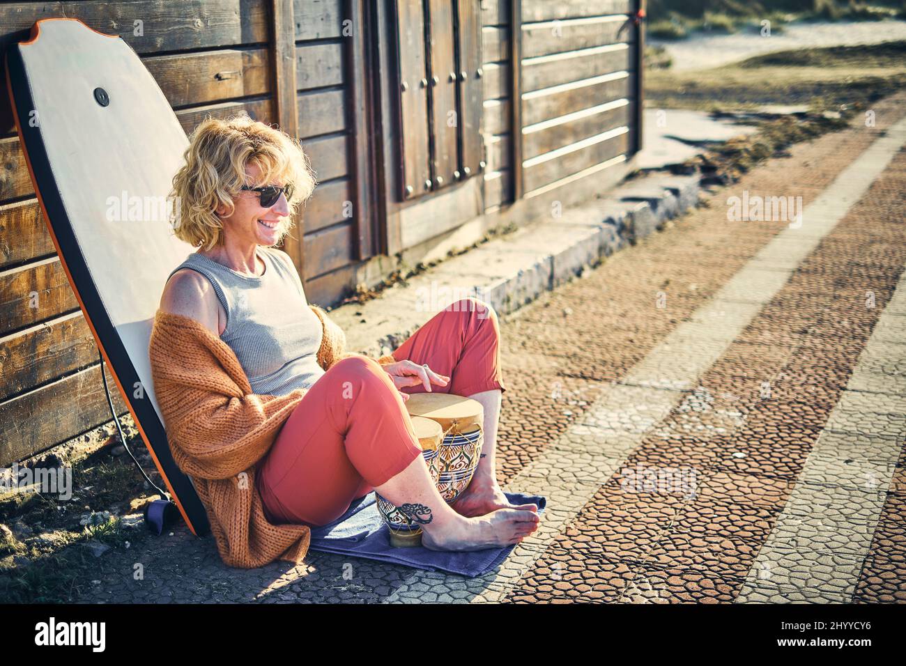 Portrait d'une jeune femme de race blanche, blonde et mûre, à l'extérieur, dans une maison en bois près d'une plage avec un bodyboard et des lunettes de soleil jouant à bongo. Berri Banque D'Images