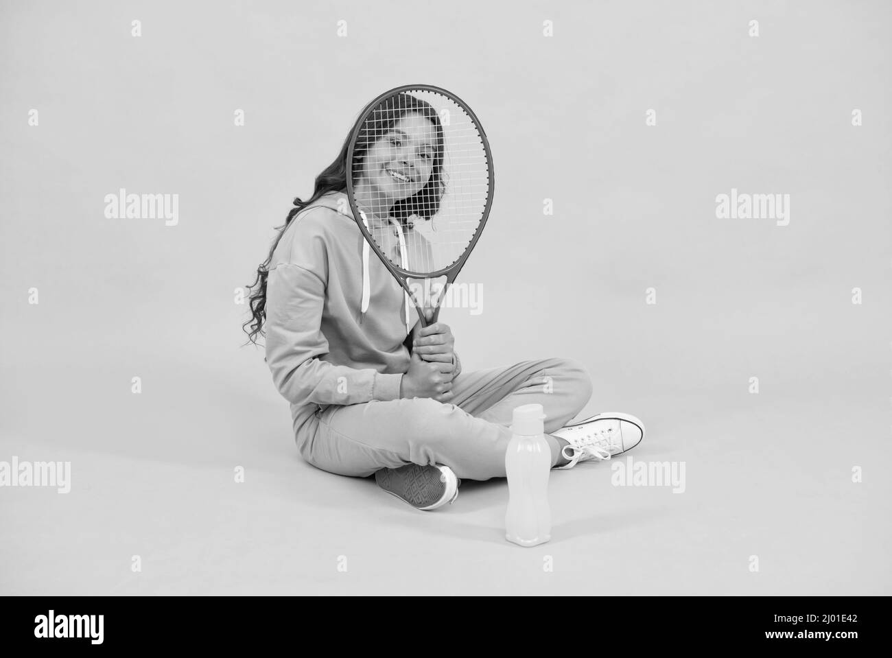 un enfant heureux s'assoit dans des vêtements de sport avec une raquette de squash et une bouteille d'eau sur fond jaune, enfant Banque D'Images