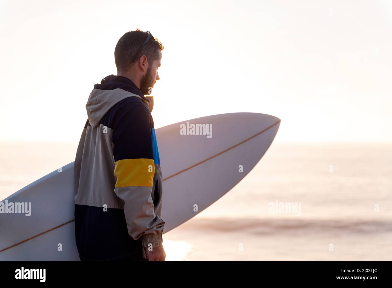 jeune homme en veste regardant la mer au coucher du soleil avec sa planche de surf sous son bras, loisirs et passe-temps concept, copier l'espace pour le texte Banque D'Images