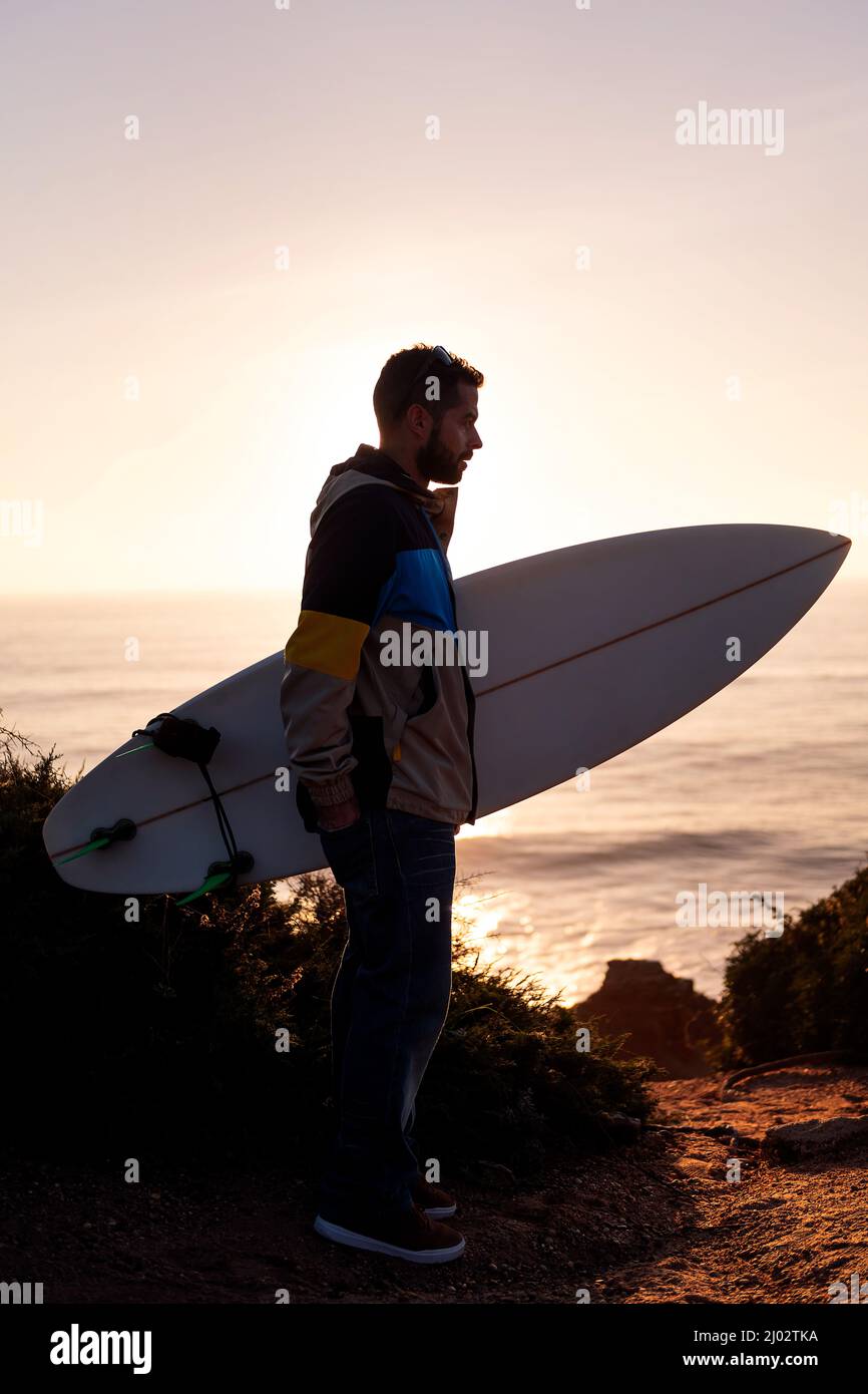 photo verticale de la silhouette d'un jeune homme dans une veste avec sa planche de surf sous son bras regardant la mer au coucher du soleil, loisirs et hobbies concept Banque D'Images