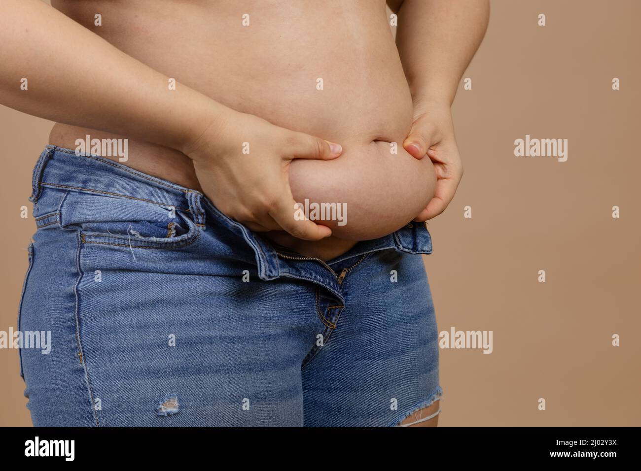 Femme tenant le gros ventre avec les mains montrant la graisse dans bleu jeans sur fond beige. Gain de poids soudain. Graisse viscérale. Corps positif. Serré peu Banque D'Images