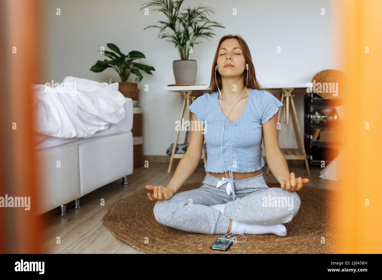 Jeune femme tendance portant des écouteurs méditant en ligne et écoutant de la musique sur l'application de lecteur de smartphone tout en étant assise au sol à la maison, application de méditation dans le cadre de la routine matinale de pleine conscience, humeur paisible. Banque D'Images