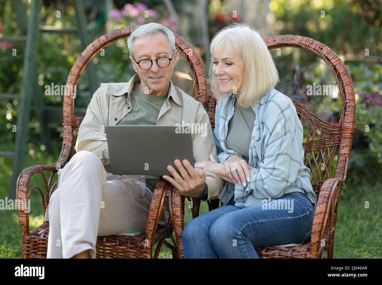 Les conjoints âgés modernes se détendent dans des chaises en osier en plein air et utilisant un ordinateur portable, en profitant de la soirée de printemps dehors Banque D'Images