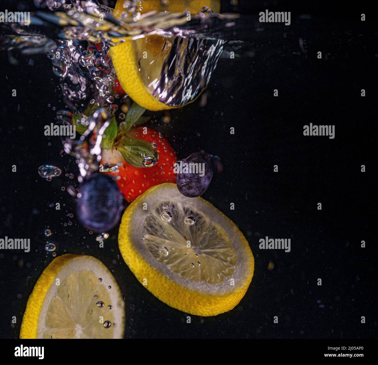 Les fraises rouges, les citrons jaunes et les bleuets tombent dans l'eau. Éclaboussures d'eau, gouttes. Mouvement lent, arrière-plan noir. Banque D'Images