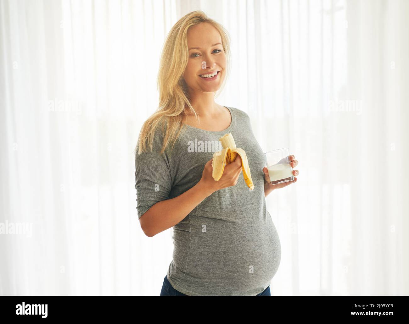 Maman en bonne santé, bébé en bonne santé. Portrait d'une femme enceinte heureuse mangeant une banane et buvant un verre de lait à la maison. Banque D'Images