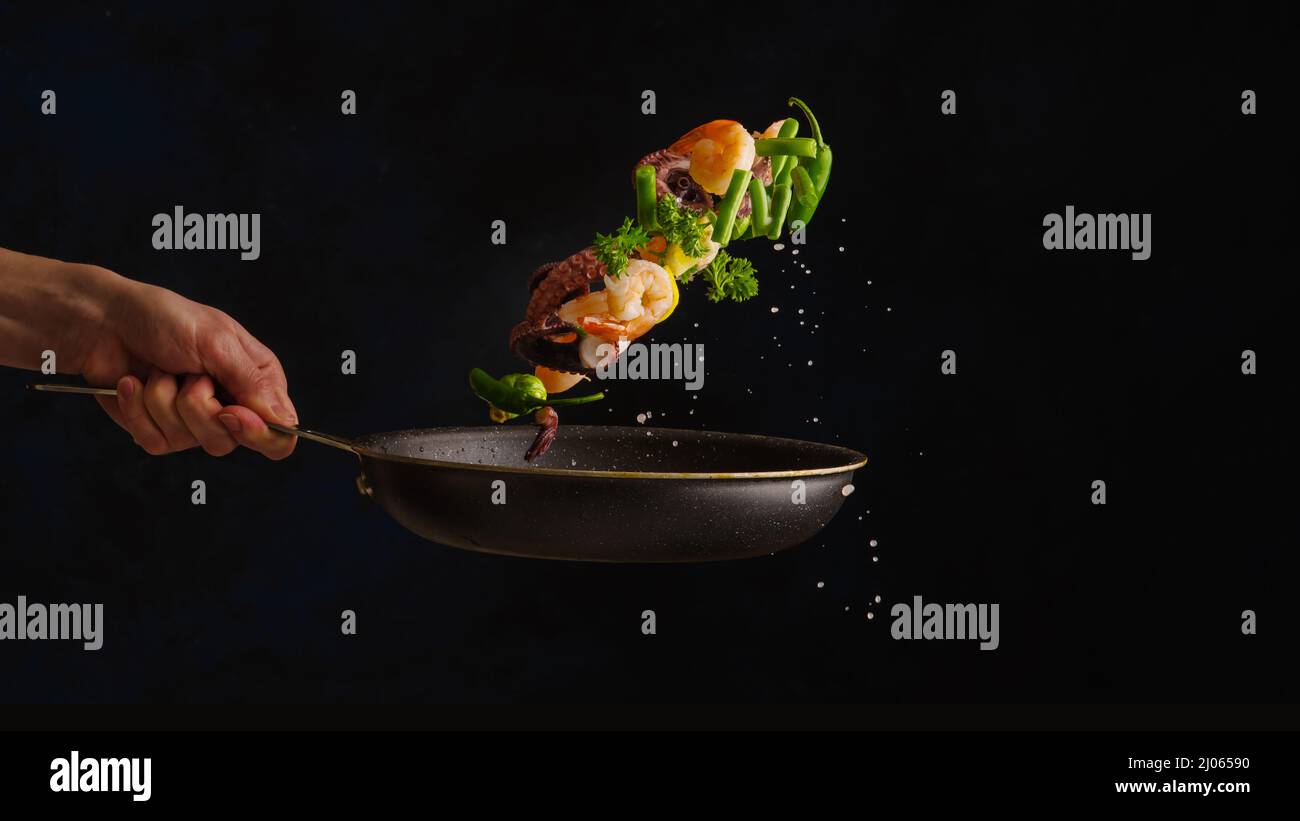 Un chef professionnel cuisine des fruits de mer - crevettes, pieuvre aux herbes et cires dans une poêle sur fond noir lors d'un vol gelé. Vue horizontale. Par Banque D'Images
