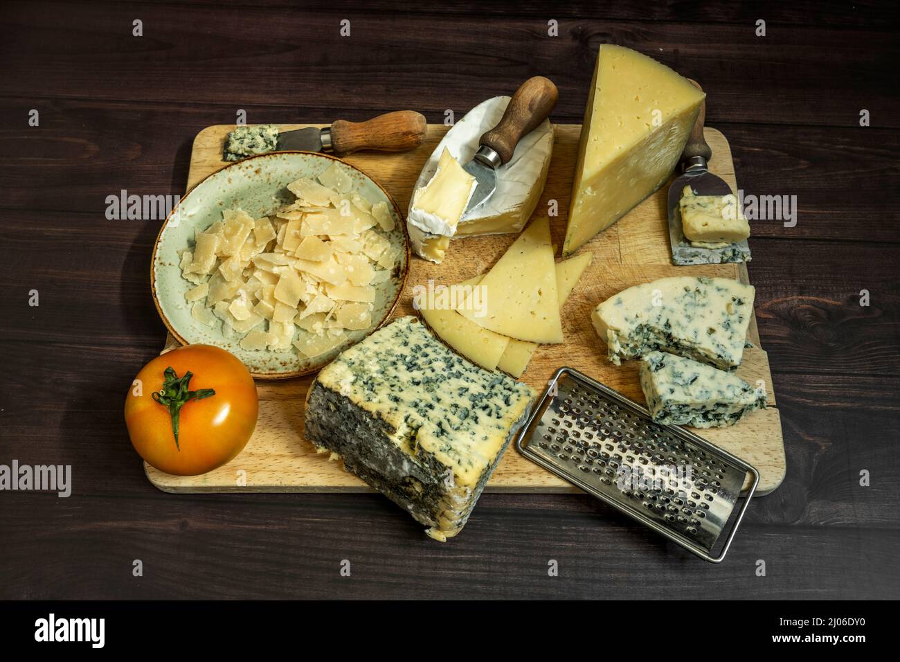 Assortiment de fromages encore en vie avec des couteaux, de la tomate, du fromage bleu, des flocons de parmesan et des grattoirs sur bois Banque D'Images