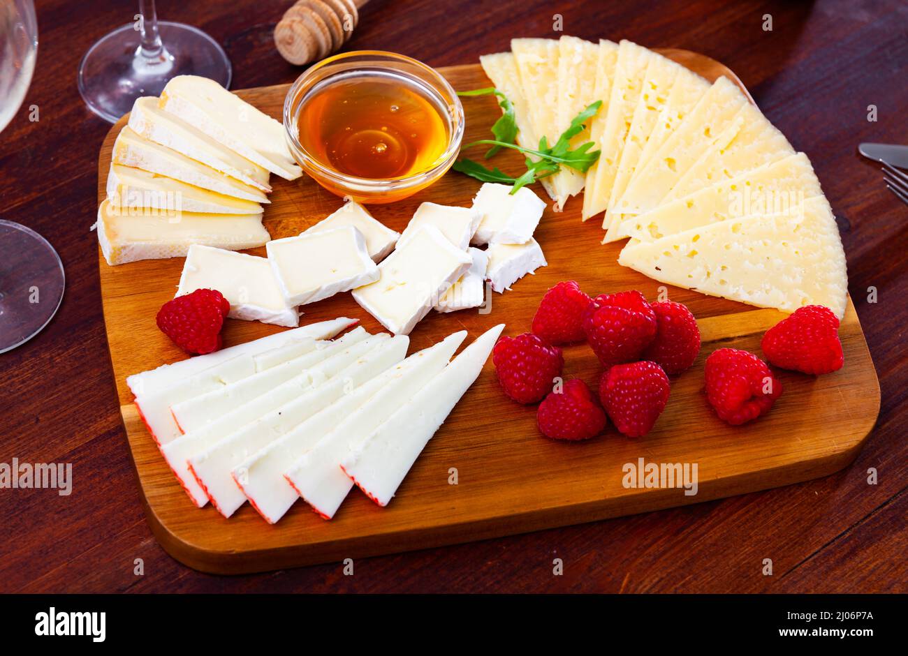Différents types de fromage tranchés et servis sur une planche de bois avec du miel et des framboises. Banque D'Images