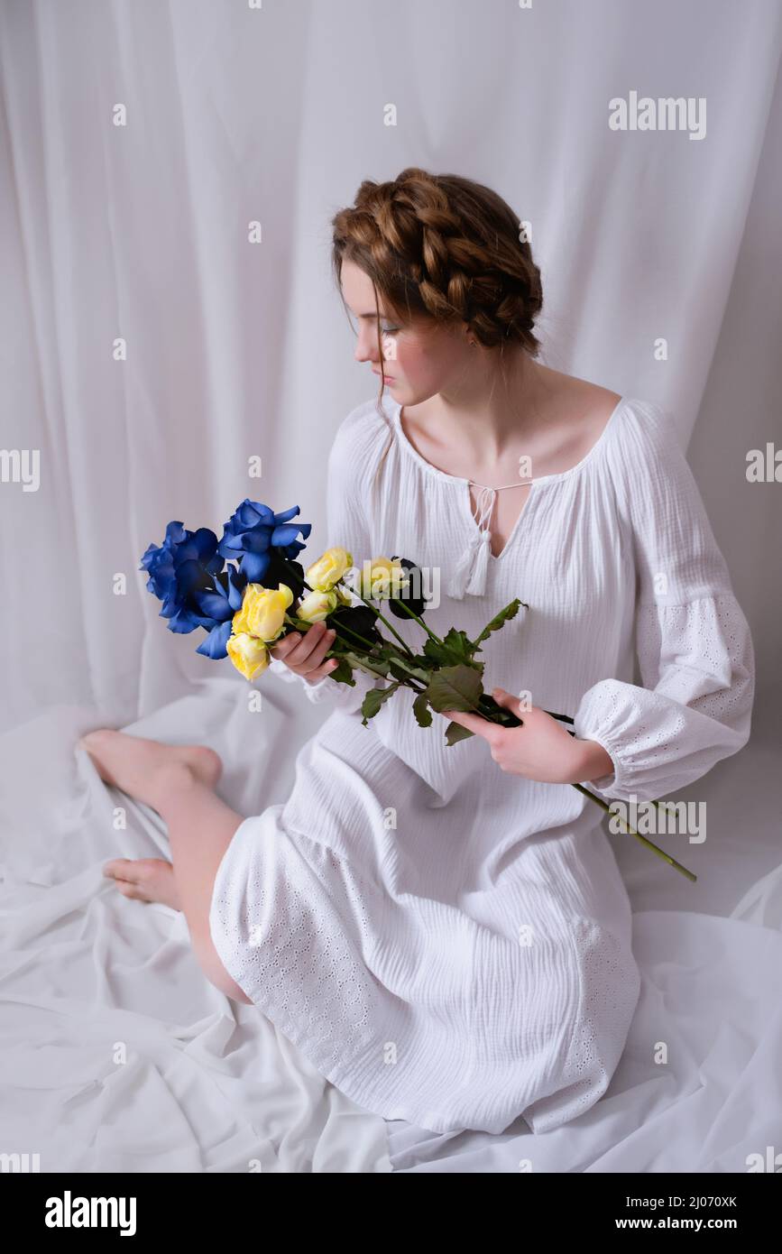 Adolescent ukrainien caucasien assis dans le studio dans une robe et des fleurs jaune-bleu sur un fond blanc . Concept - mettre fin à la guerre en Ukraine. Un ukrainien libre, une nation invincible Banque D'Images