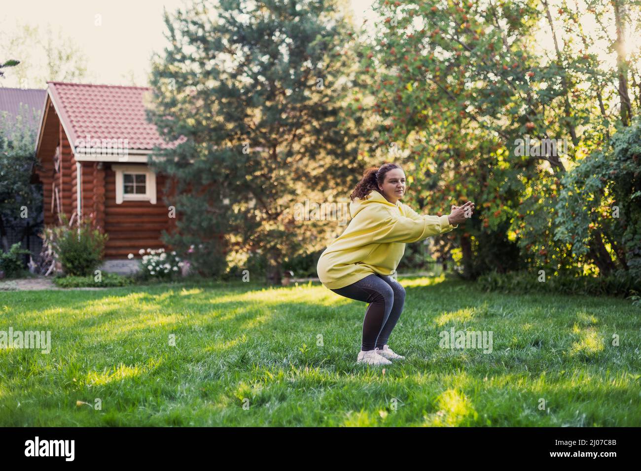 Femme sur l'herbe verte faisant le yoga squating dans les vêtements de sport regardant la caméra sur l'arrière-cour plein de lumière du soleil avec maison de campagne et de hauts arbres dedans Banque D'Images