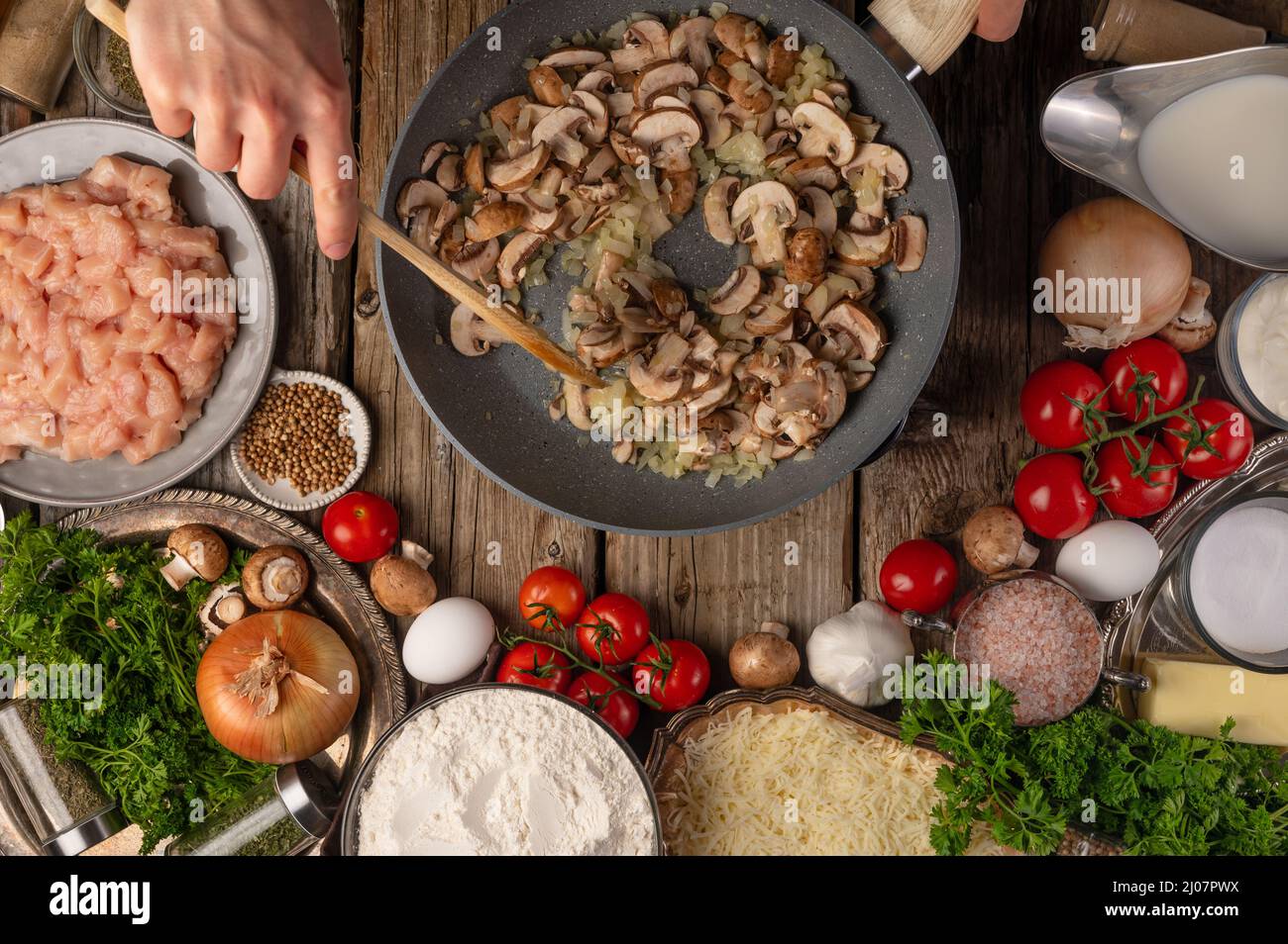 Les mains du chef remuent les champignons dans la poêle pour préparer le remplissage sur une table en bois avec une variété d'ingrédients de fond. Concept du processus de cuisson. Banque D'Images