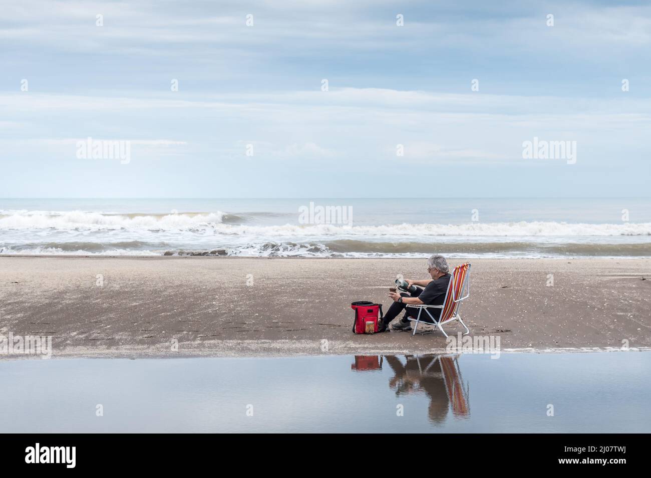 Homme adulte mature assis sur une chaise de plage versant de l'eau dans un compagnon tout reflété dans l'eau et dans le fond les vagues de la mer. Banque D'Images