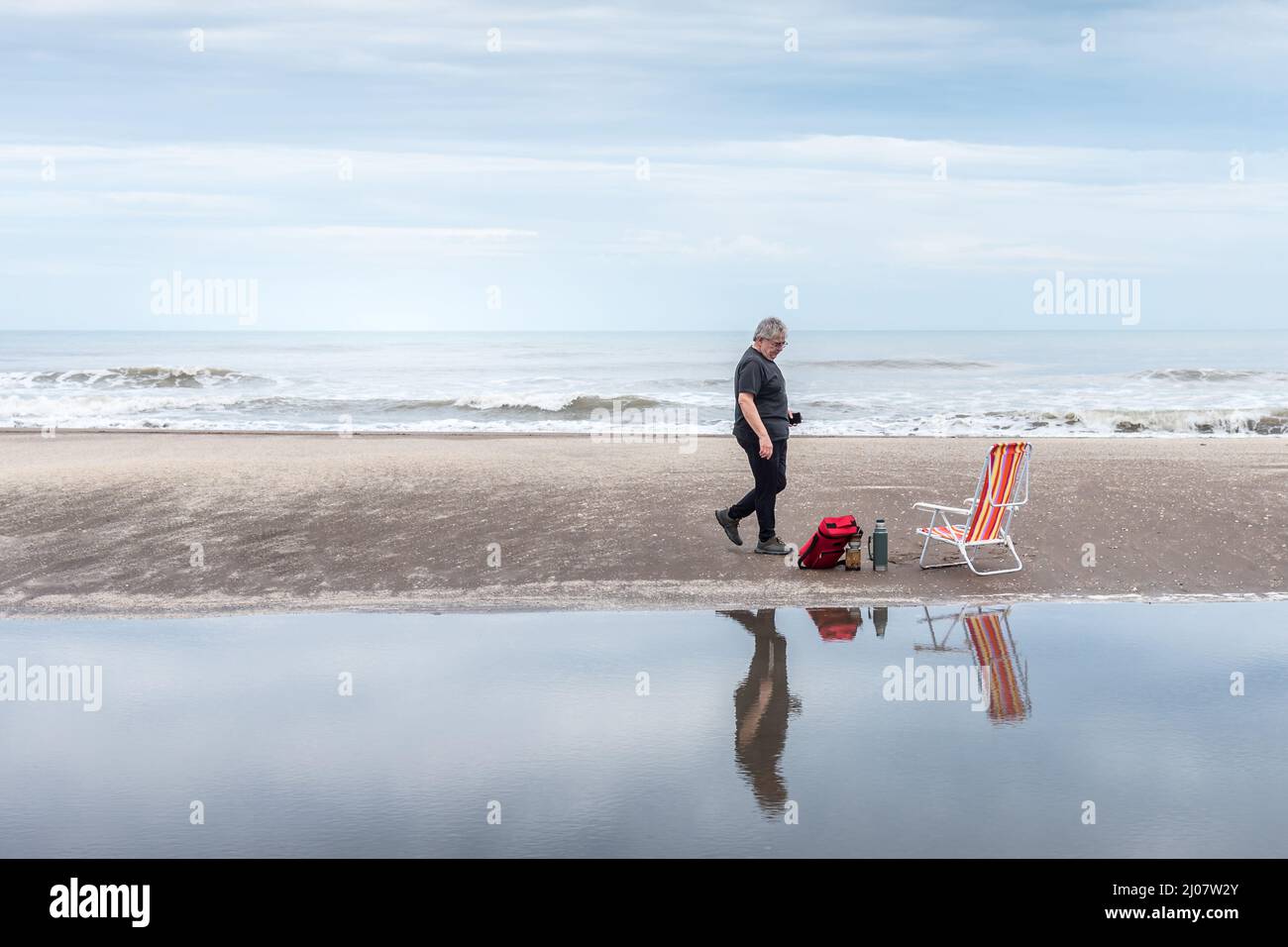Homme mûr marchant le long de la plage vers un ensemble de maté à côté d'une chaise de plage reflétée dans l'eau et dans le fond les vagues de la mer. Banque D'Images